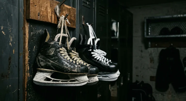 Two pairs of hockey skates hanging from a locker room hook — one worn and scuffed, one brand new — moody sports lighting